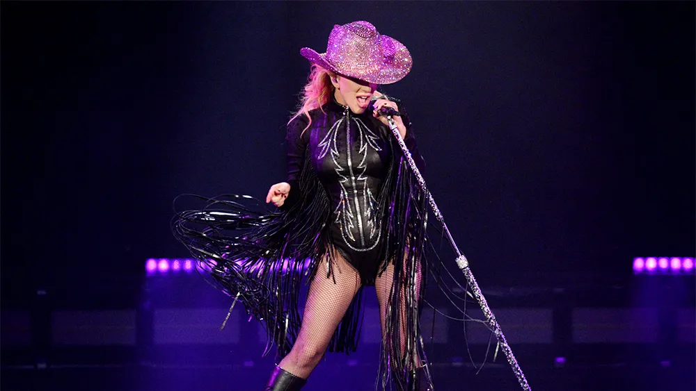 Lady gaga pop star performing on stage in a black fringed outfit and glittering pink cowboy hat, holding a sparkling microphone stand under dramatic lighting.