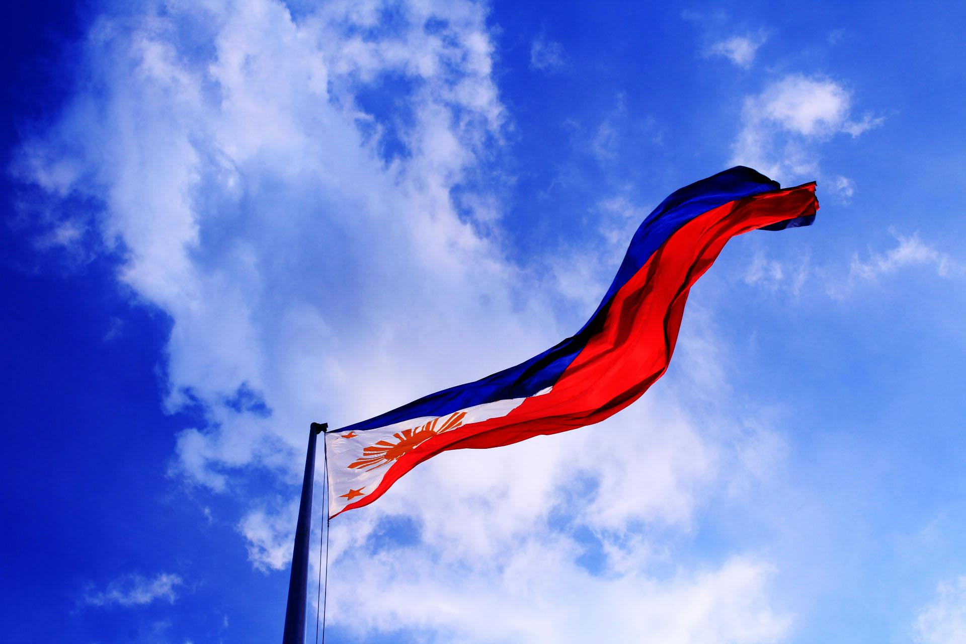 Philippine national flag waving against a bright blue sky with scattered clouds, symbolizing patriotism and national pride.