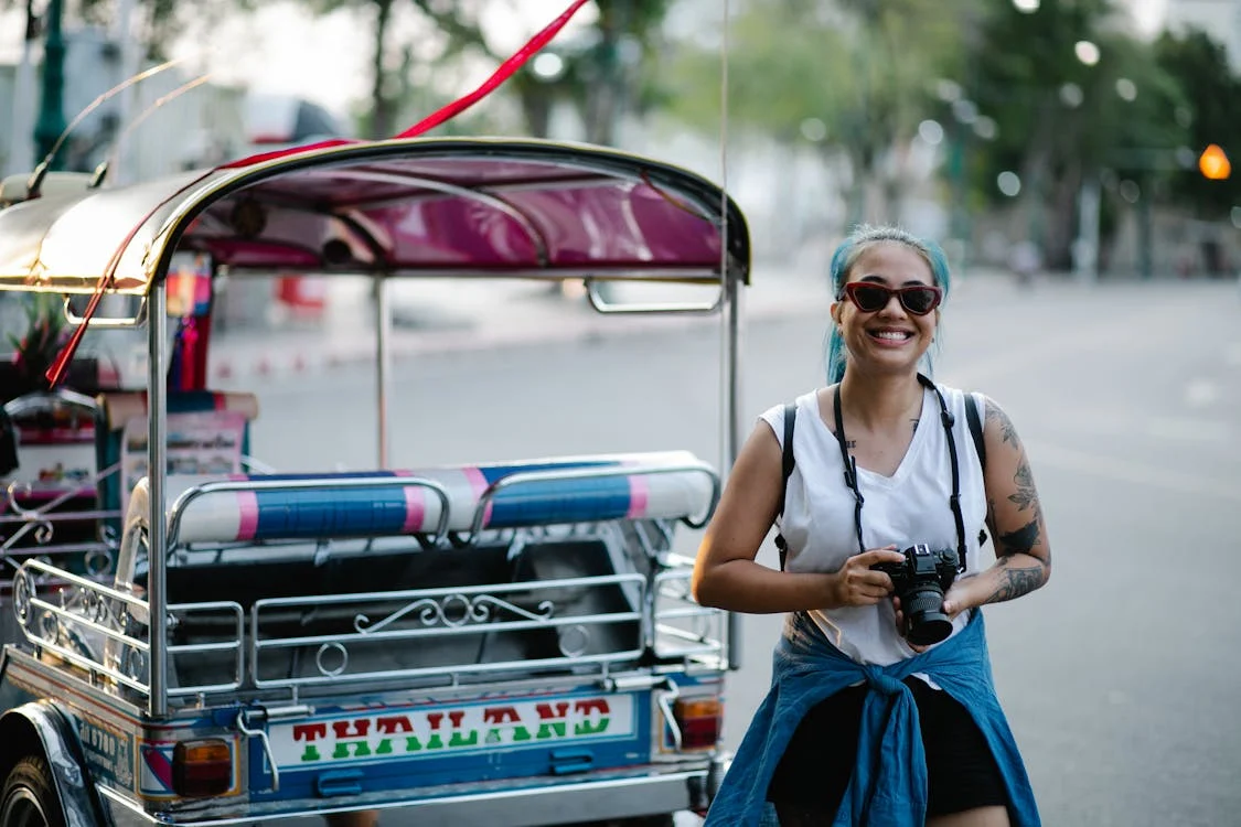 Smiling tourist with camera next to colorful tuk-tuk on street in Thailand.