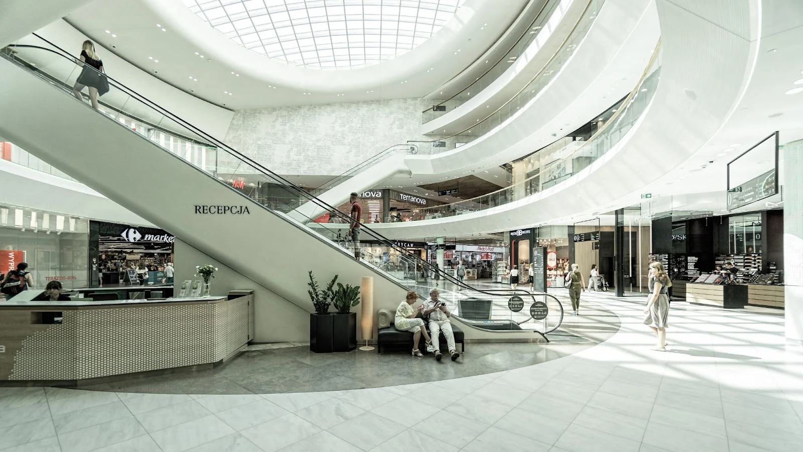 Modern shopping mall interior with curved architecture, natural lighting from a glass dome ceiling, and international retail stores like Carrefour and Terranova, reflecting contemporary European retail design.