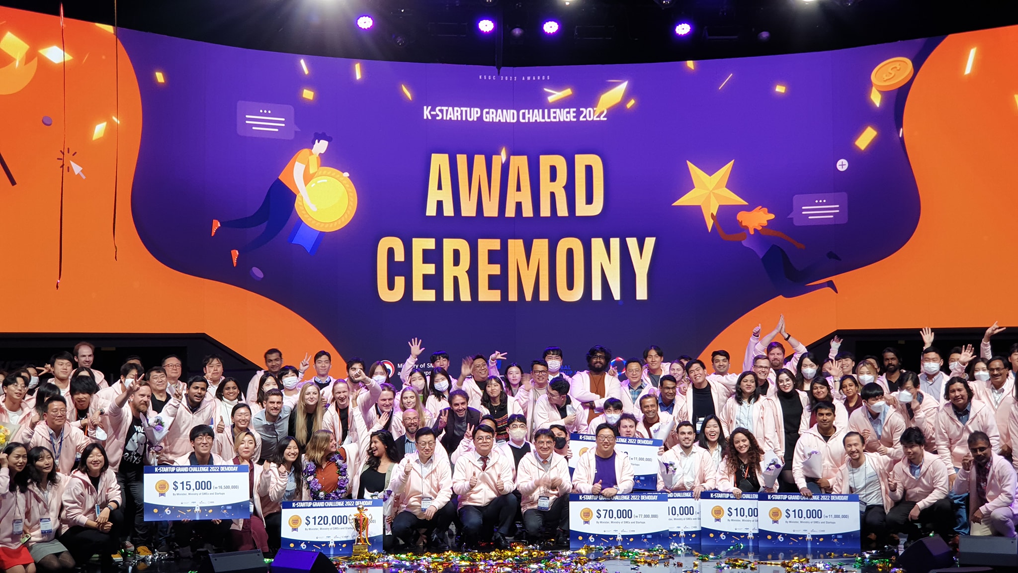 A large group of participants and winners celebrate on stage at the K-Startup Grand Challenge 2022 Award Ceremony. The vibrant backdrop features bold text reading “AWARD CEREMONY” with animated illustrations of achievers. Attendees are dressed in pink jackets, smiling and holding giant cheques with various cash prizes, surrounded by confetti and celebratory energy.
