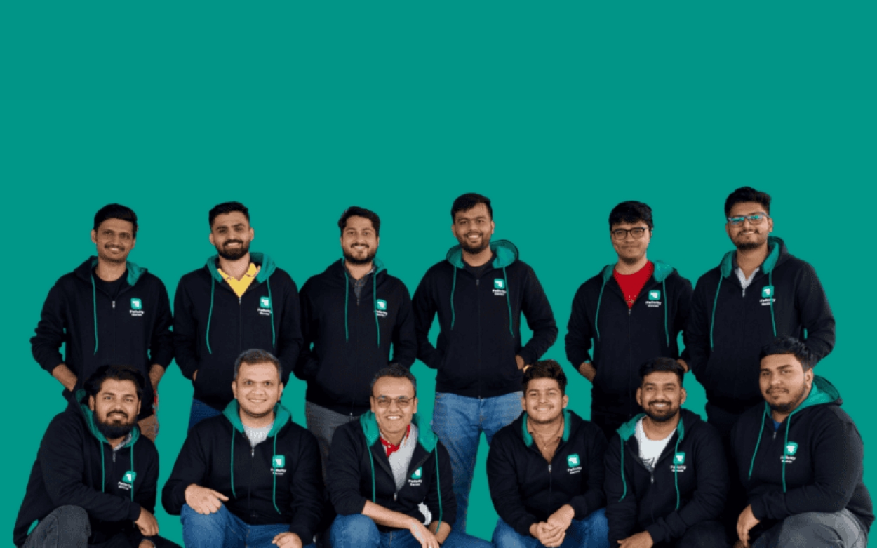 Group photo of thirteen team members wearing matching black hoodies with a green and white company logo, posing in front of a solid teal background.