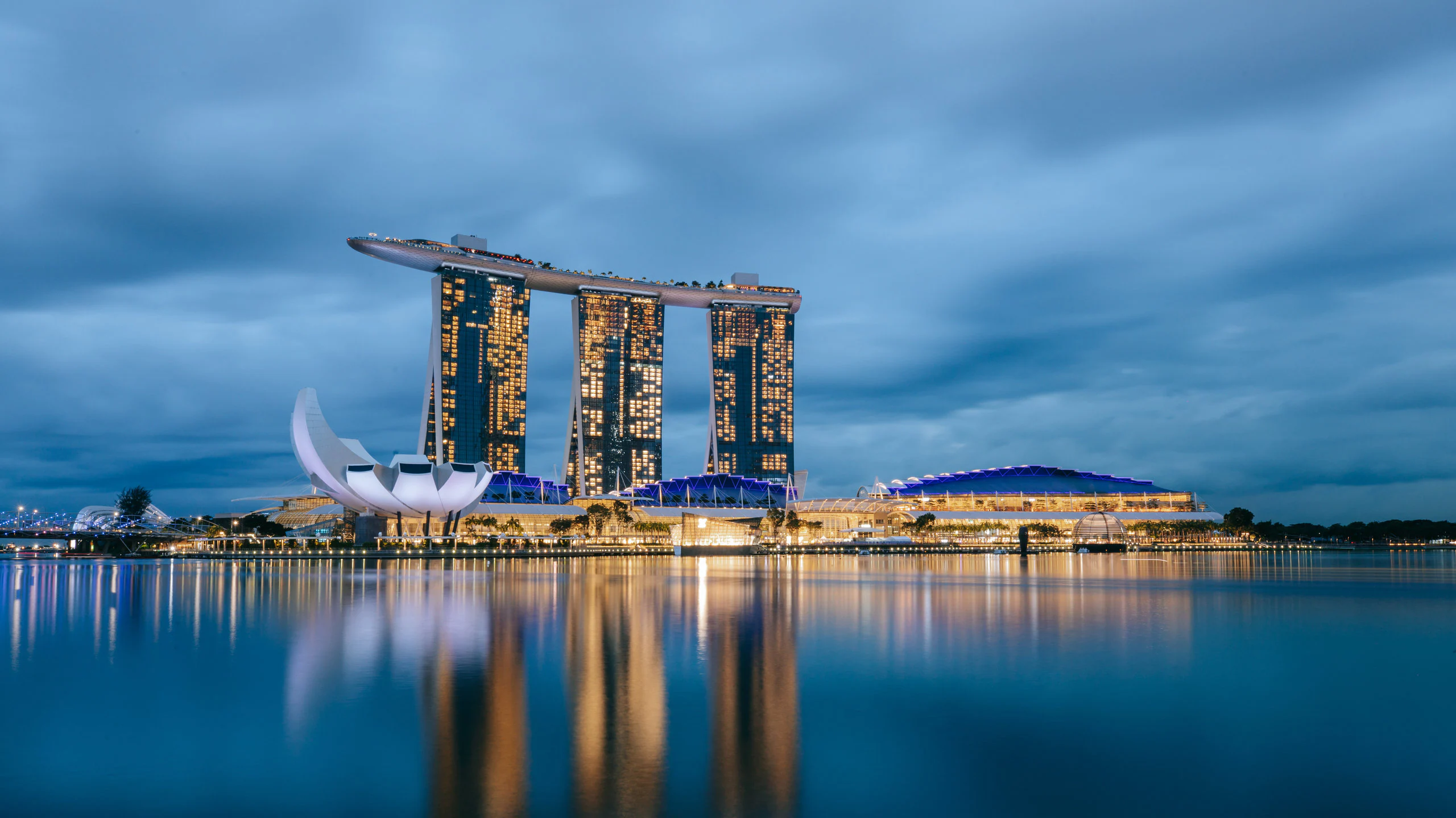 Night view of Marina Bay Sands in Singapore, with its three illuminated towers and rooftop SkyPark reflected on the calm Marina Bay waterfront under a cloudy blue evening sky.