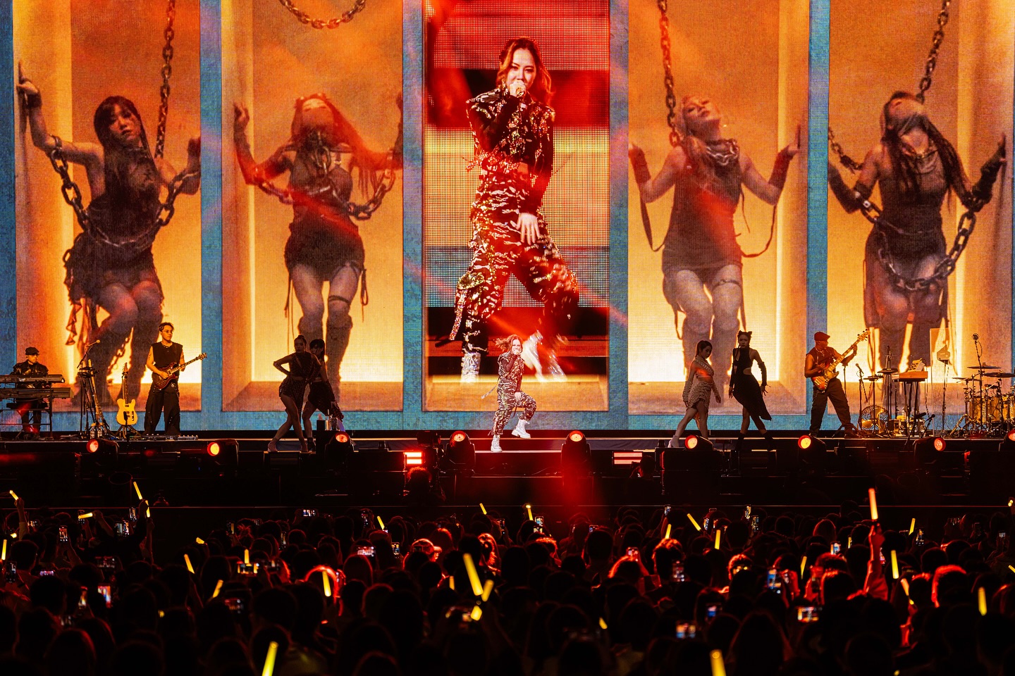 Live concert performance with dynamic stage visuals, female lead singer, backup dancers, and cheering crowd with glow sticks.