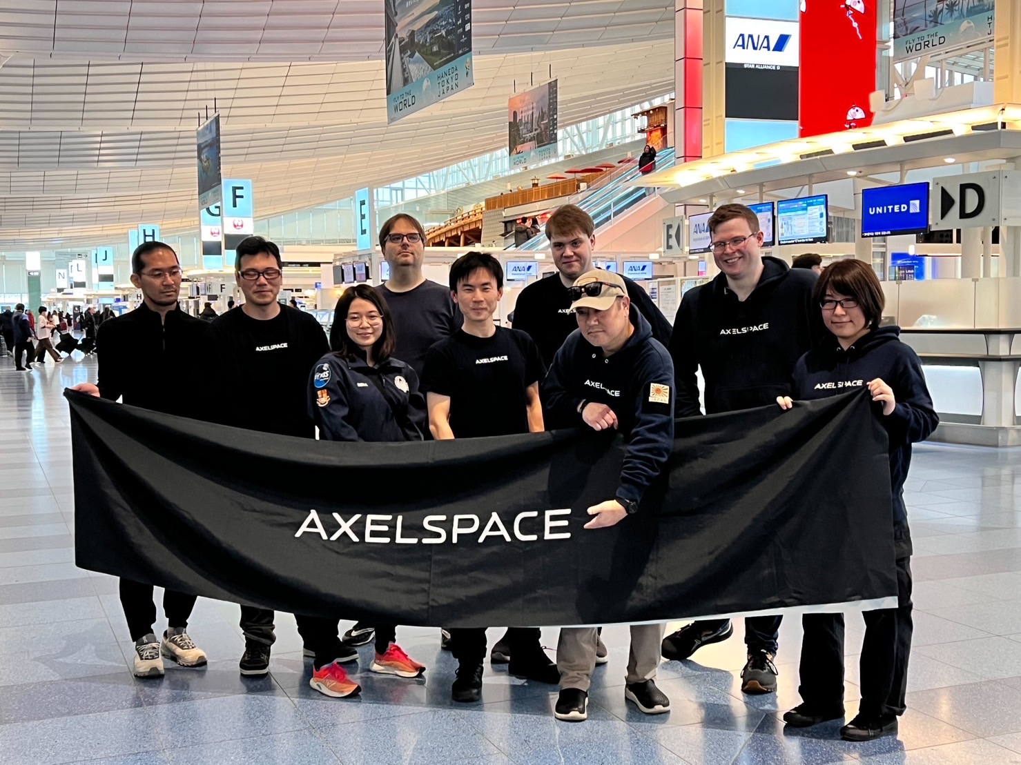 Axelspace team posing with a company banner at an airport terminal, showcasing international collaboration in aerospace technology.