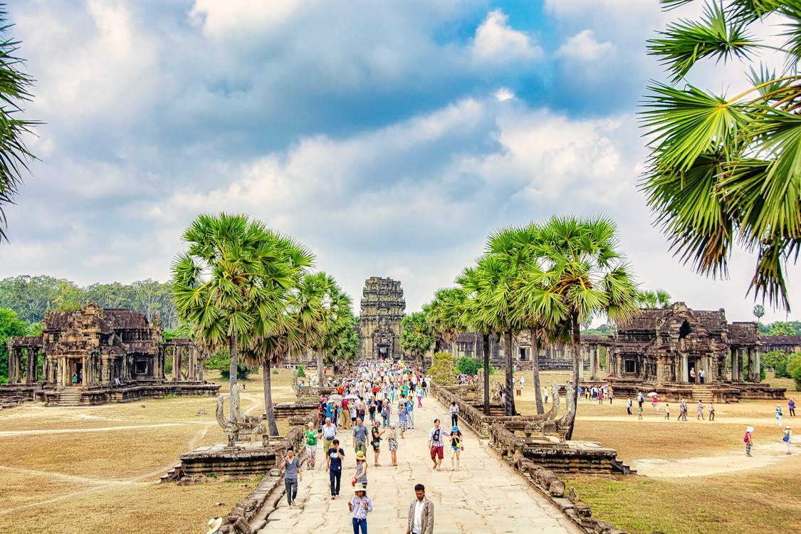 Tourists exploring the ancient Angkor Wat temple complex in Cambodia, surrounded by palm trees and historic stone architecture under a partly cloudy sky.
