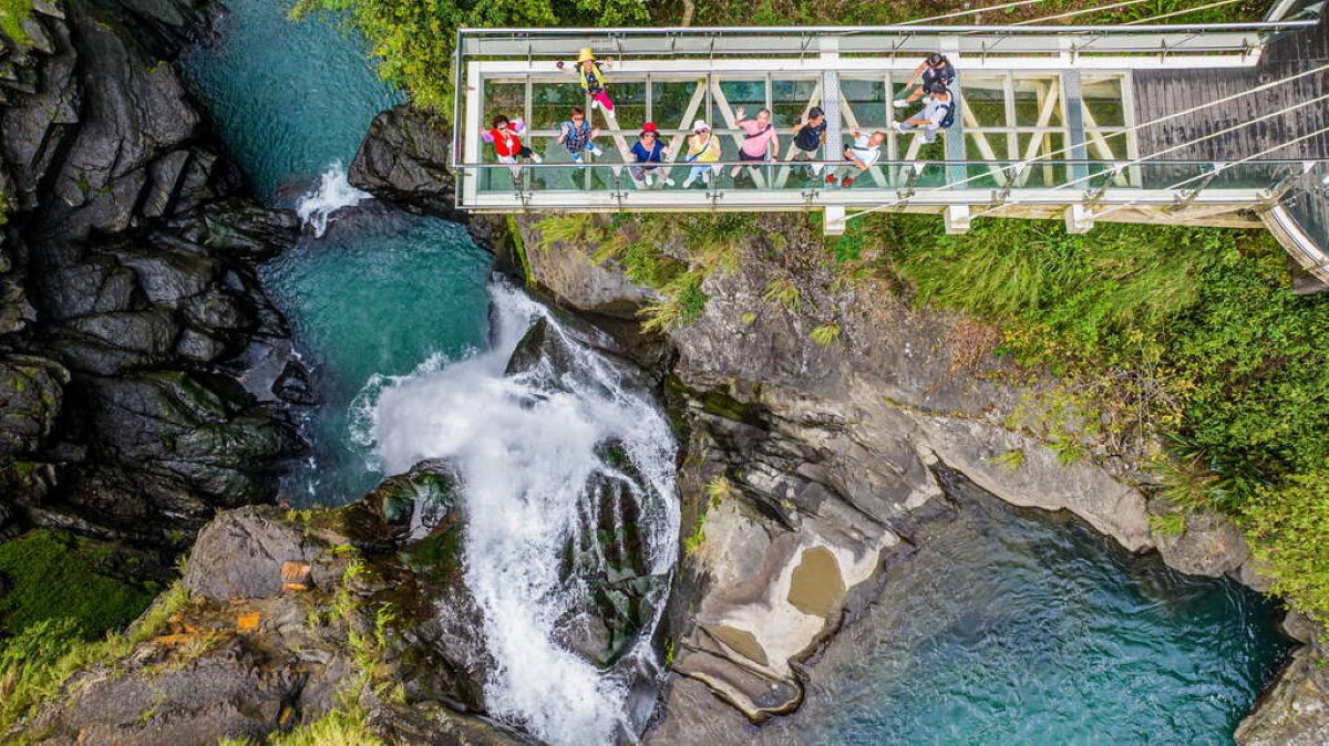 Overhead view of tourists on a glass suspension bridge above a scenic waterfall and turquoise stream in a forested canyon, showcasing eco-tourism and adventure travel.
