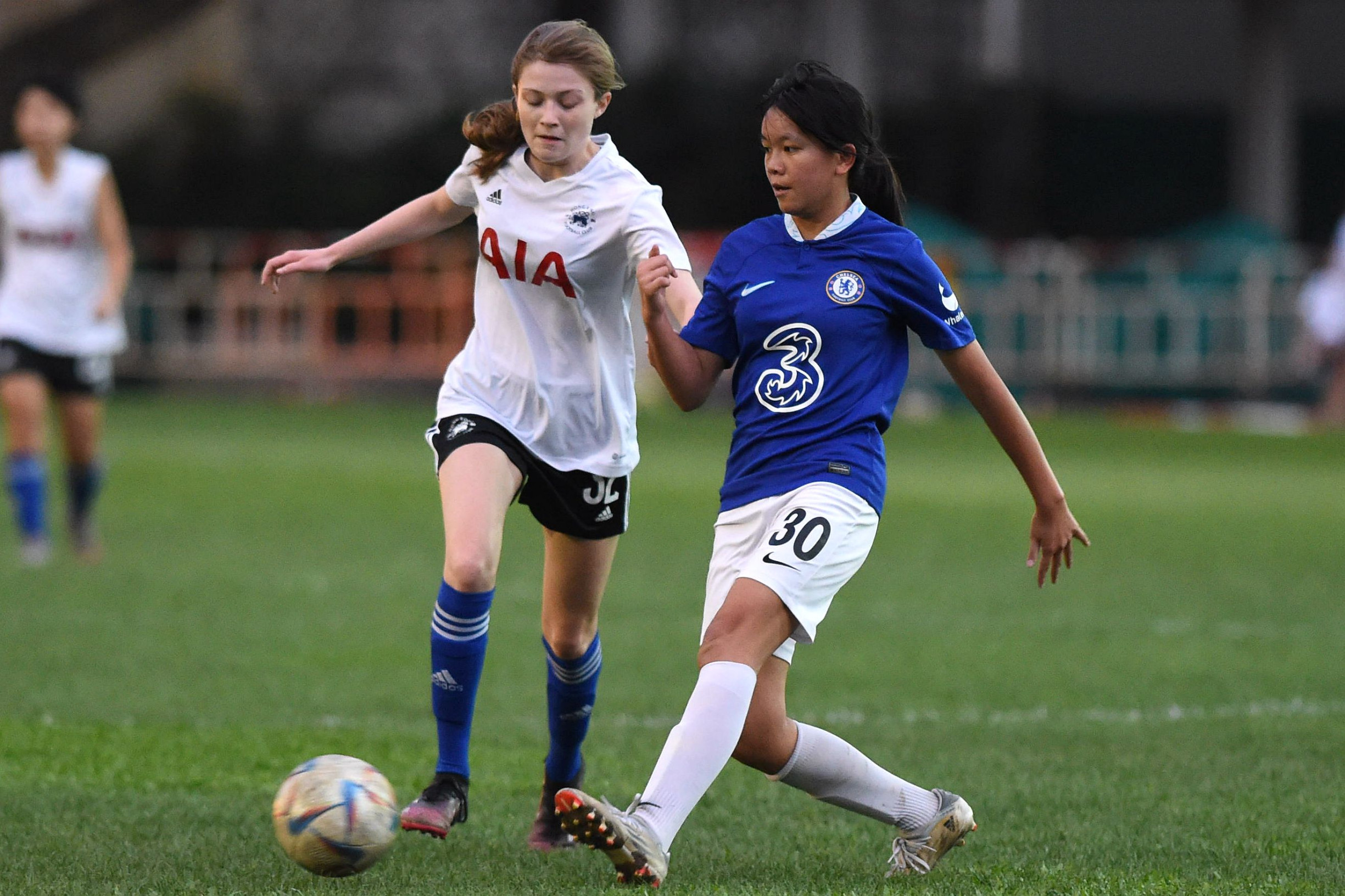 Two young female soccer players compete for the ball during a match, one wearing a Chelsea FC blue kit and the other in a Tottenham Hotspur white jersey, on a grassy field.