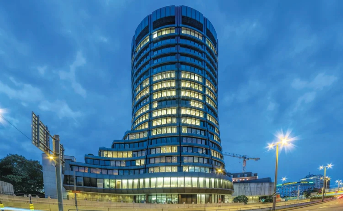 The illuminated headquarters of the Bank for International Settlements in Basel, Switzerland, captured at dusk with a vibrant cityscape background.