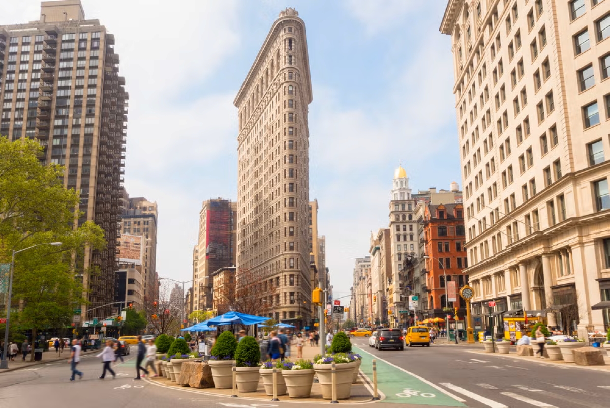 Street-level view of the Flatiron Building in New York City on a sunny day, with pedestrians, cars, and outdoor dining setups creating a vibrant urban scene.