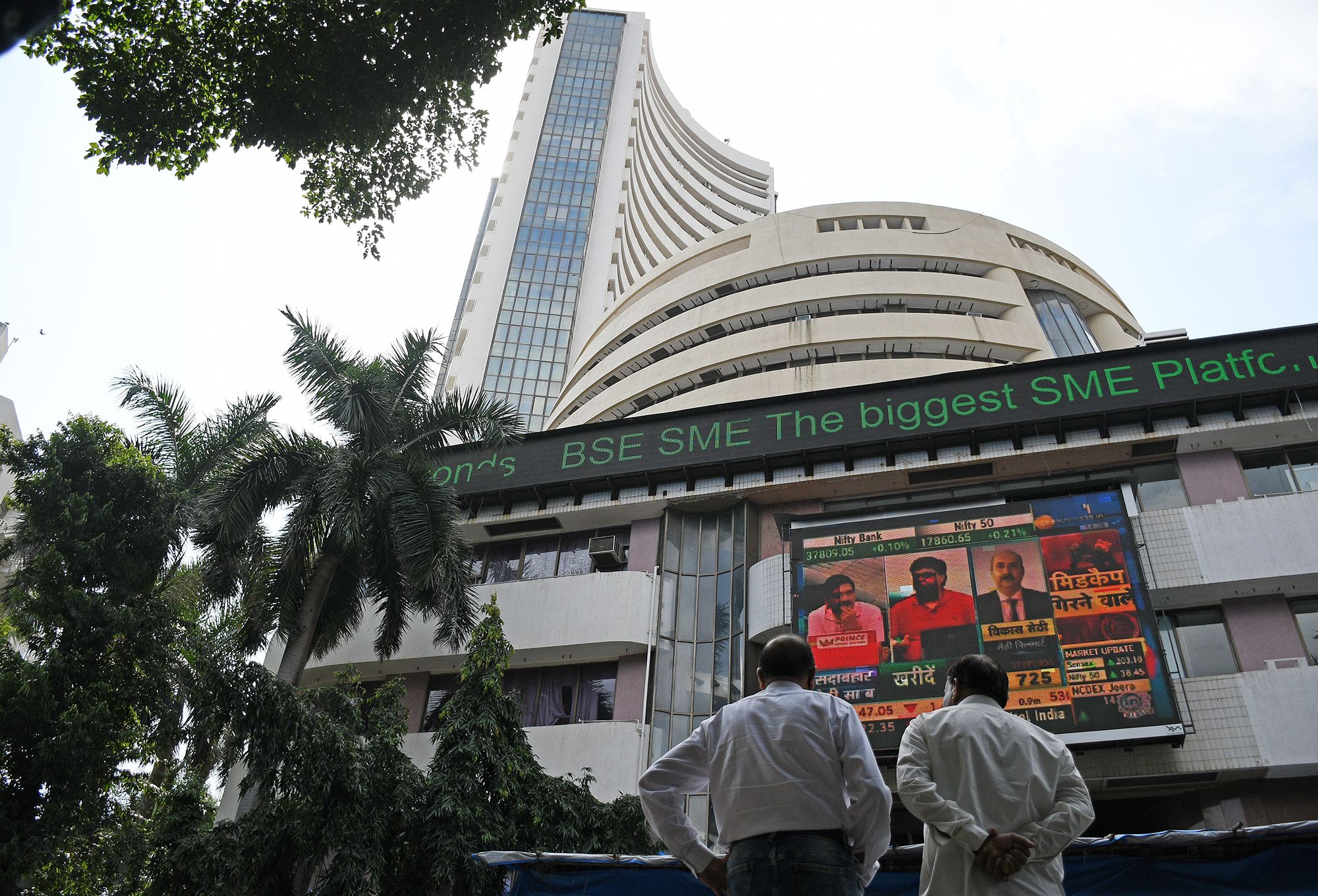 People standing in front of the Bombay Stock Exchange (BSE) building in Mumbai, India, looking at a large digital screen displaying stock market updates and news.