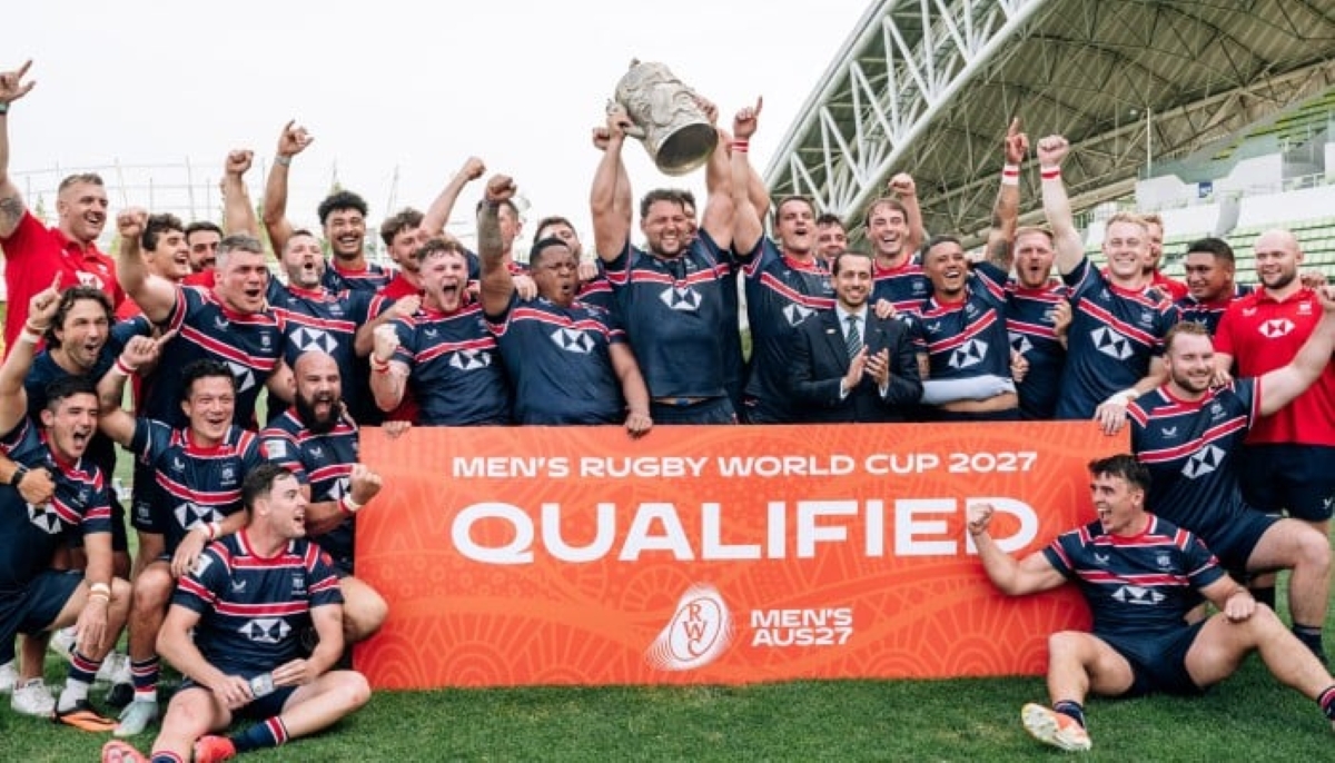 The USA men’s rugby team celebrates qualification for the Men’s Rugby World Cup 2027, holding a trophy aloft and posing behind an orange banner that reads "QUALIFIED." The players and coaching staff are dressed in matching navy blue kits with red and white accents. Their expressions show joy and triumph as they pose on a stadium field with a modern, arched roof structure in the background.