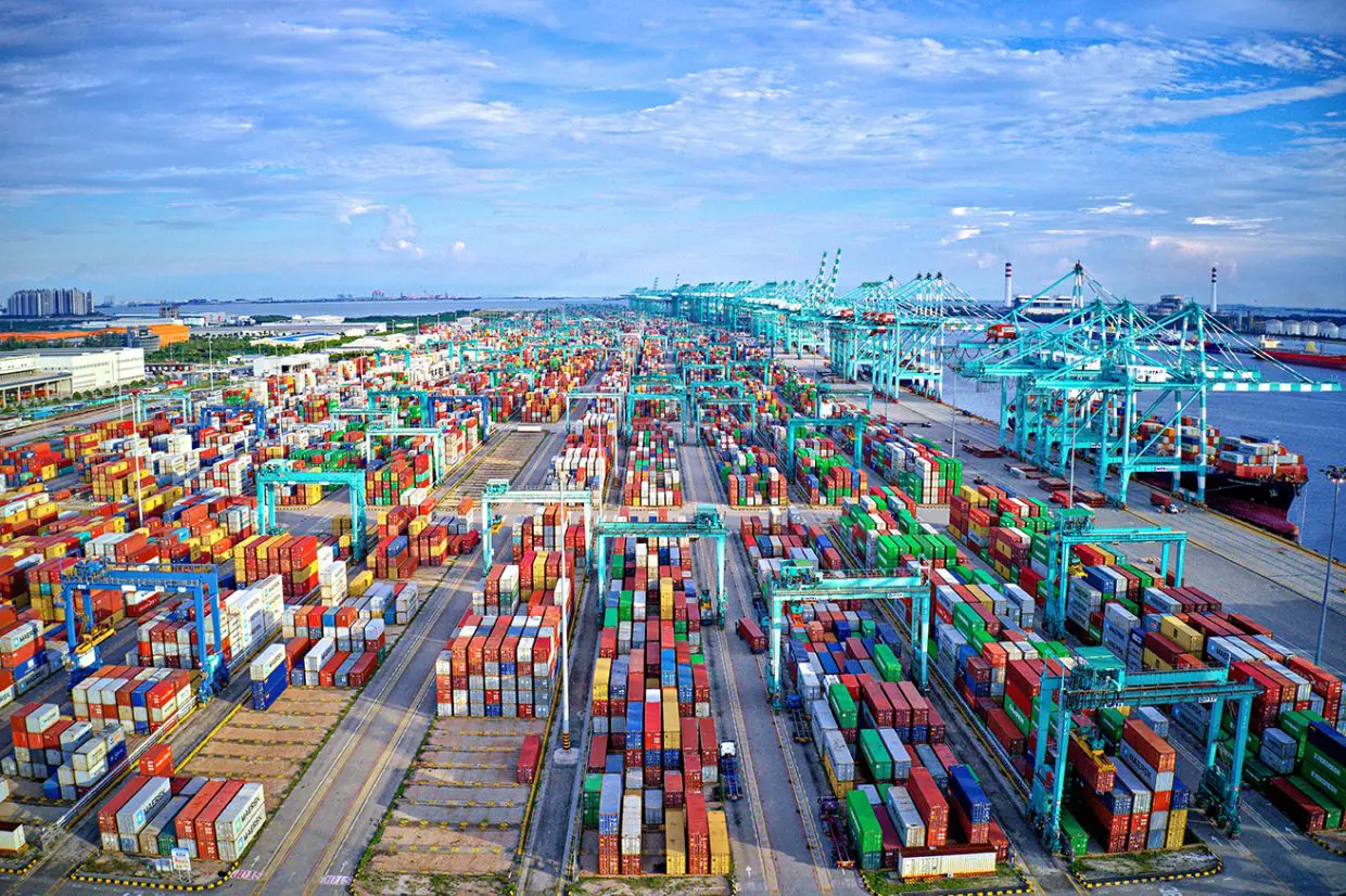 Aerial view of a major Asian container port with colorful shipping containers, automated cranes, and docked cargo ships under a clear blue sky.