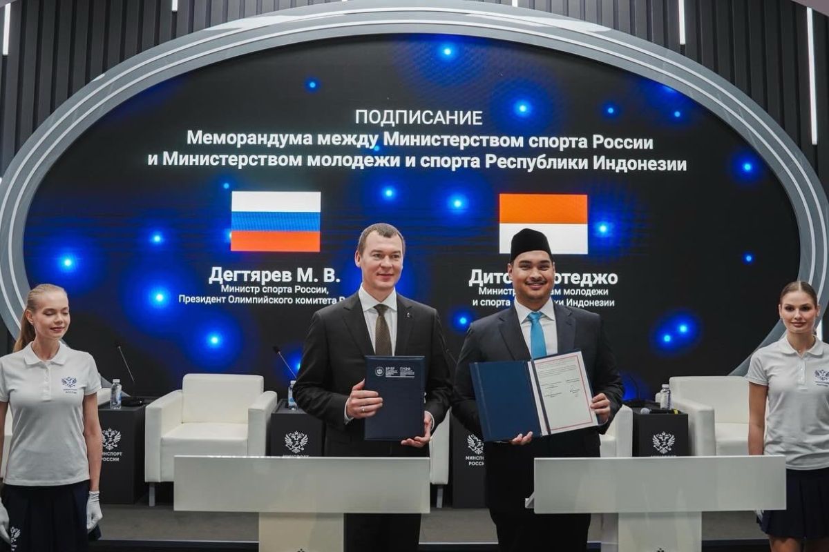 A smiling track athlete in racing gear poses in a victory stance reminiscent of Usain Bolt, pointing both arms diagonally upward. He is standing on a red running track, wearing a bib number 427. In the background are mountains, sports facilities, and cameras set up on tripods, suggesting a competitive event in progress.