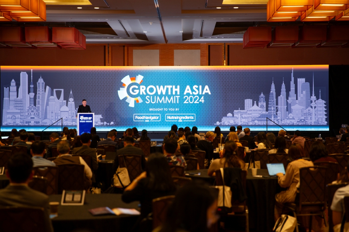 A large audience attends the Growth Asia Summit 2024, held in a grand conference hall. The stage backdrop prominently displays the event’s title alongside stylized city skylines representing key Asian cities. A speaker stands at the podium addressing attendees, while people sit at round tables, some with laptops and notebooks open. The summit is organized by FoodNavigator Asia and NutraIngredients Asia, as indicated on the banner. The ambiance is professional, reflecting a high-profile regional business or innovation conference.