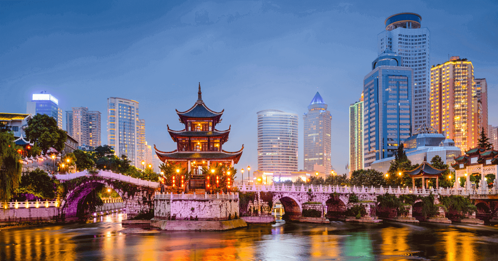 A serene evening view of Jiaxiu Pavilion in Guiyang, China, beautifully illuminated and surrounded by the Nanming River. The historic pagoda stands in contrast to the modern city skyline of tall buildings, creating a striking blend of traditional Chinese architecture and urban development. Reflections from lights dance on the river’s surface under the twilight sky.