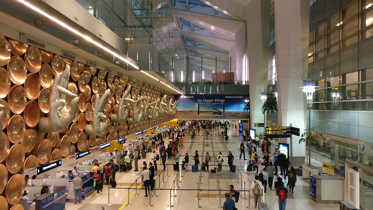 Busy terminal at Indira Gandhi International Airport in Delhi, featuring a large wall installation of mudras (hand gestures), check-in counters, and international travelers queuing under modern architecture.