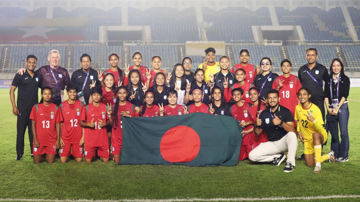 The Bangladesh women's national football team poses proudly on the field for a group photo after a match. The players and coaching staff, dressed in red and black kits, smile and flash peace signs while holding up the Bangladeshi flag in the front row. The stadium's empty seats and bright floodlights form the background, capturing a celebratory and victorious atmosphere.