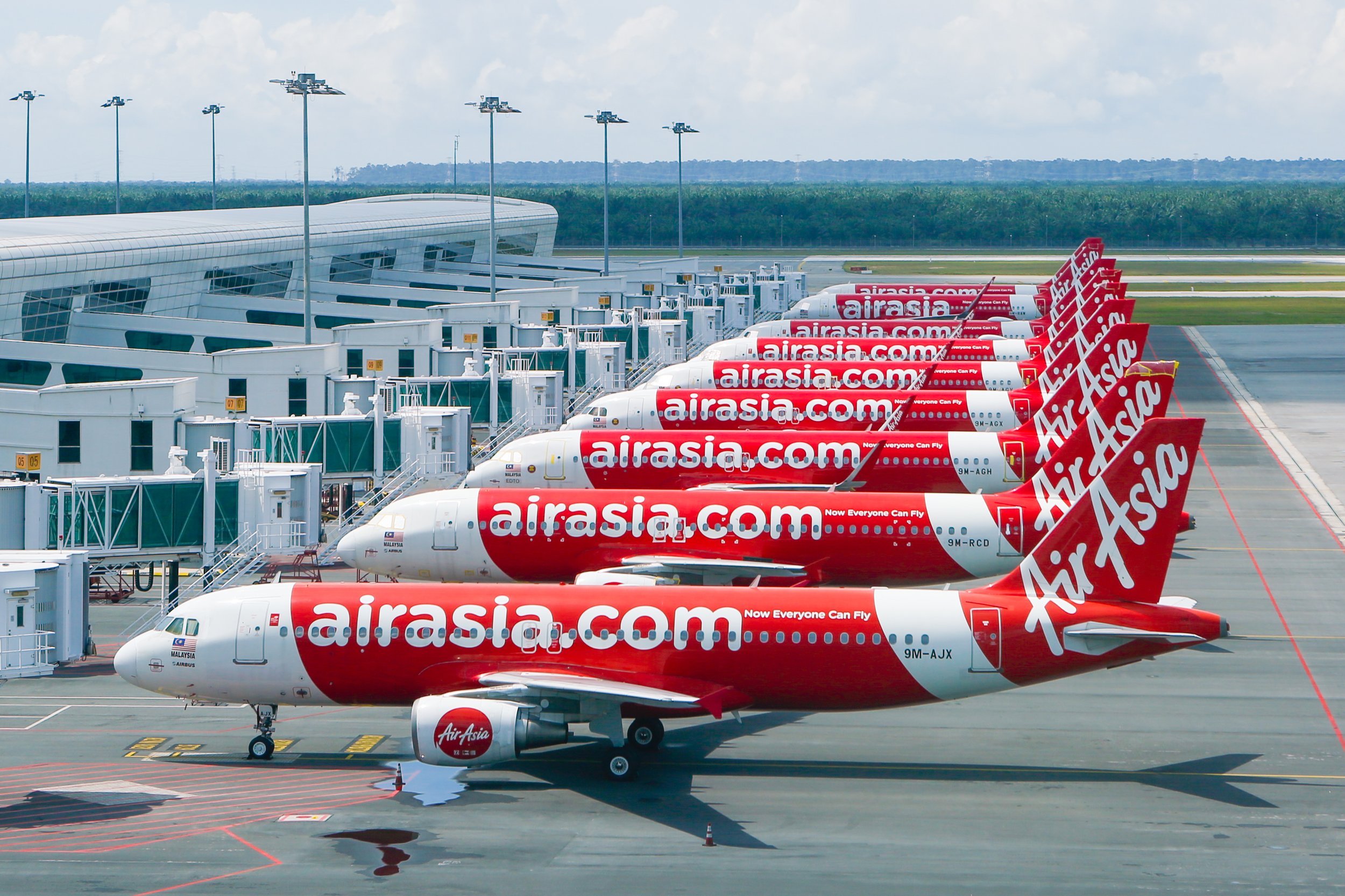A fleet of AirAsia airplanes lined up at airport gates, featuring the airline’s signature red-and-white livery with “airasia.com” boldly displayed across the fuselage. The aircraft are parked at a modern terminal with jet bridges connected, ready for boarding or deboarding. The scene captures a sense of operational scale and uniformity, symbolizing the airline’s regional dominance in low-cost air travel.