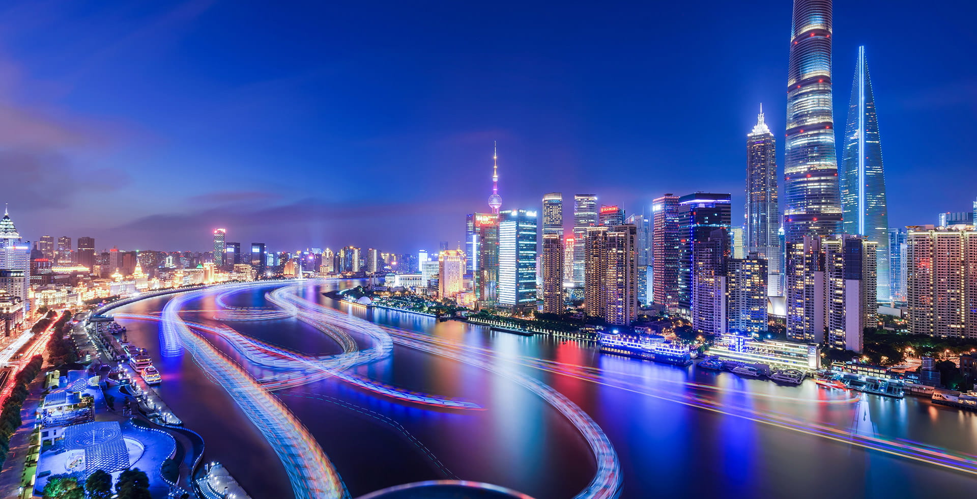 Nighttime panoramic view of Shanghai’s Lujiazui skyline with light trails from boats along the Huangpu River and glowing city skyscrapers.