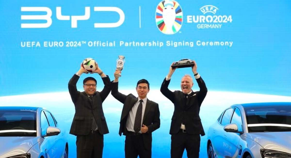 BYD and UEFA executives celebrate their EURO 2024 official partnership at a signing ceremony, holding a football, trophy, and car model in front of branded electric vehicles.