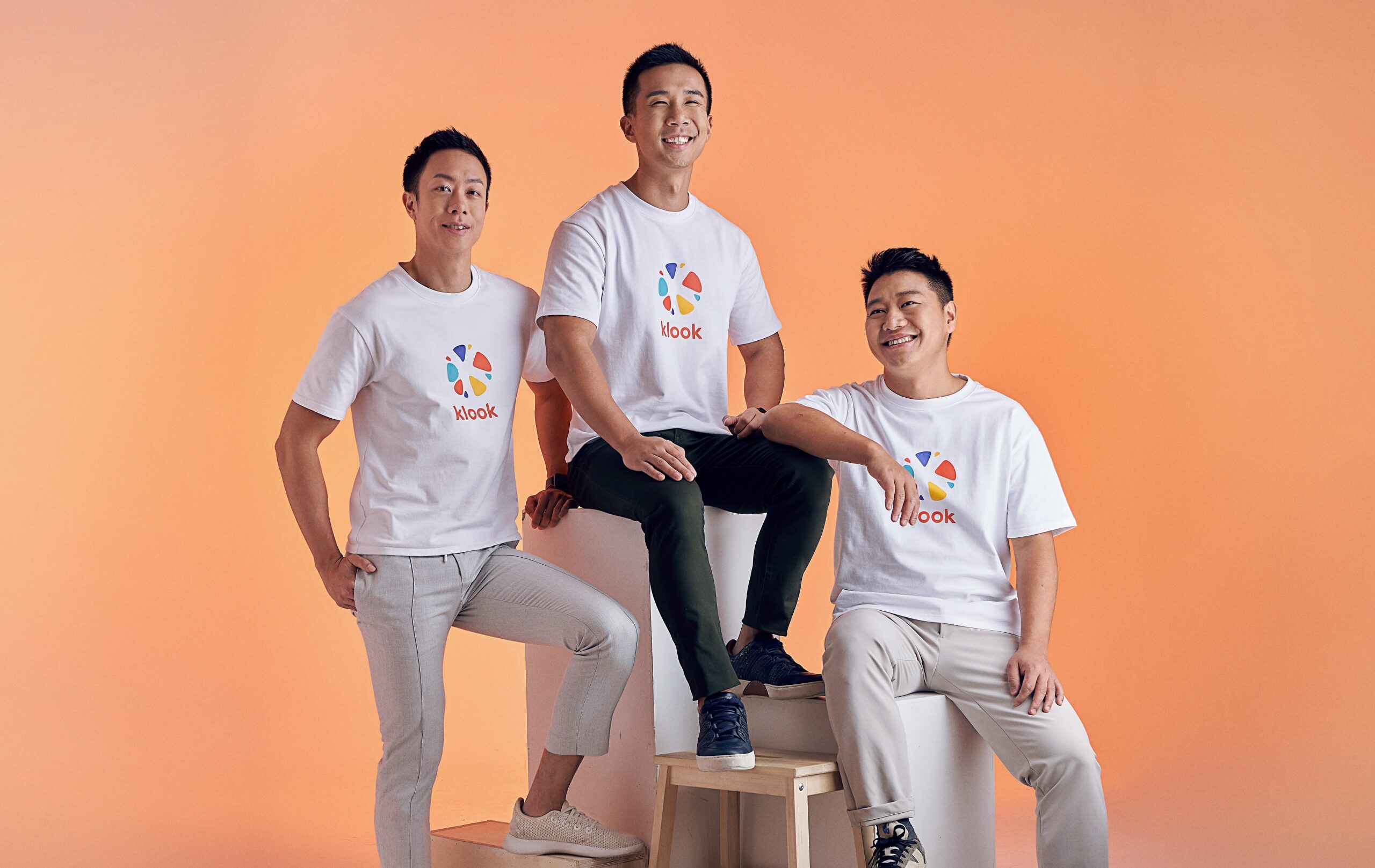 Klook co-founders posing together in branded white T-shirts against an orange backdrop, representing the travel tech company’s leadership team.