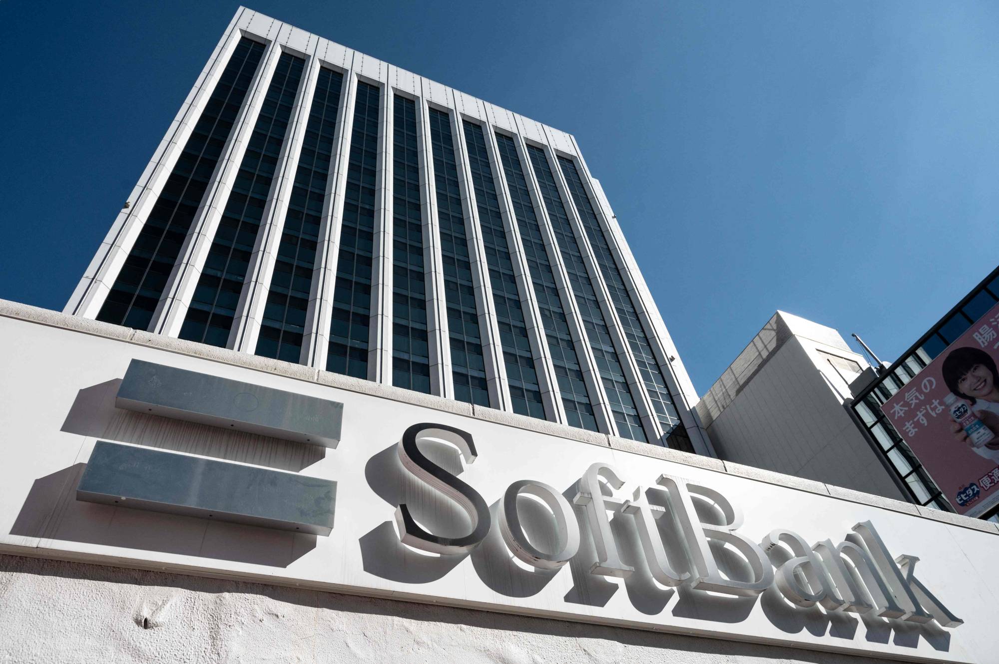 SoftBank headquarters building with company logo displayed on the exterior wall in front of a tall office tower under clear blue sky.