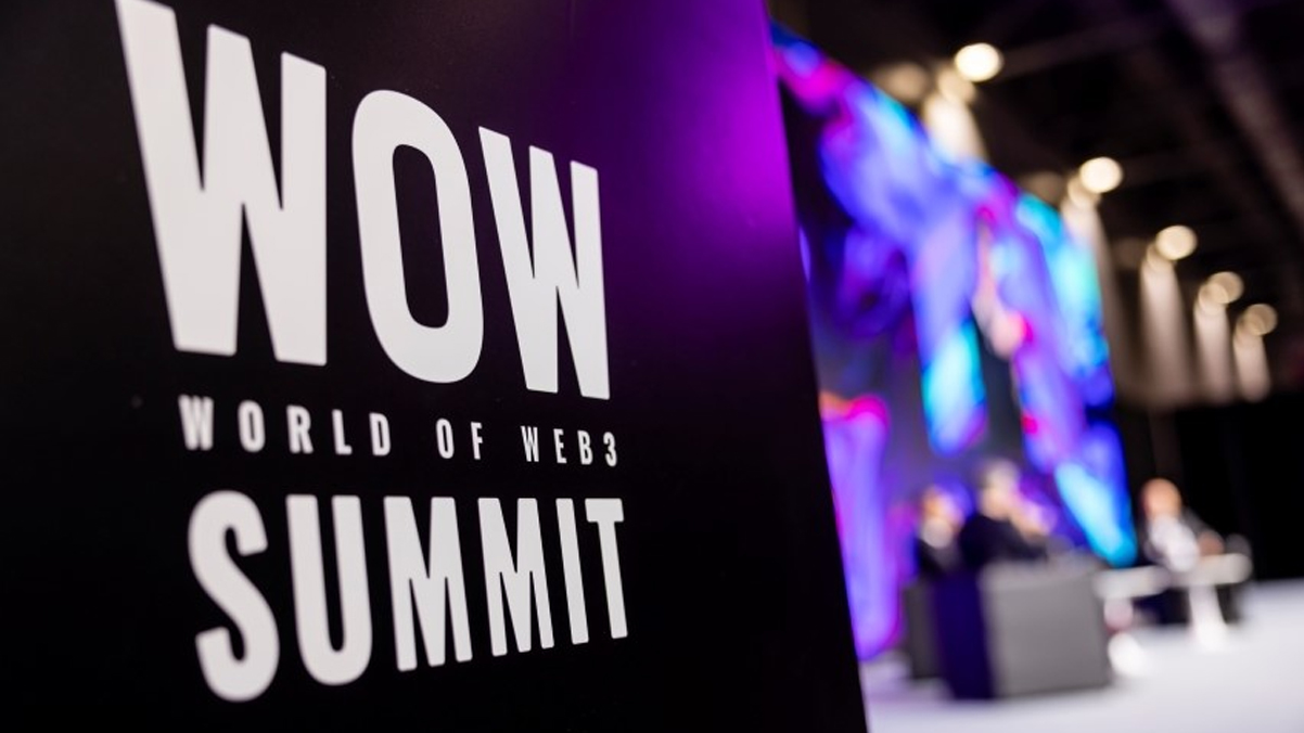 WOW – World of Web3 Summit” sign in the foreground with a blurred panel discussion stage in the background, lit by vibrant purple and blue lights.