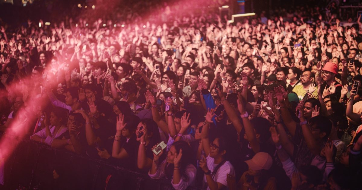 Excited crowd at a live music festival, with fans raising their hands and capturing the moment on phones under vibrant stage lighting.