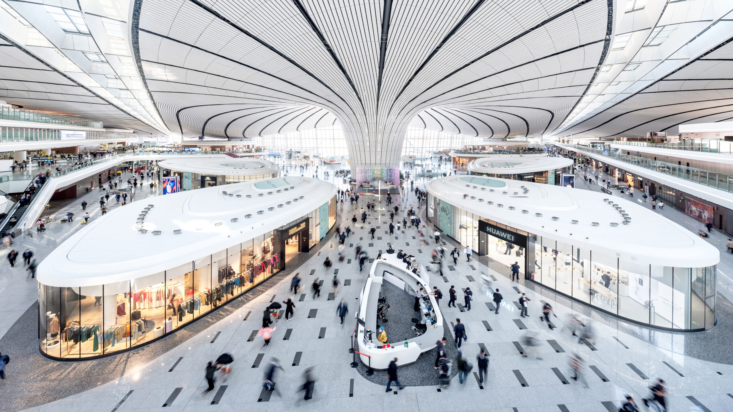Interior view of Beijing Daxing International Airport with modern architecture, retail stores, and passengers moving through the terminal.