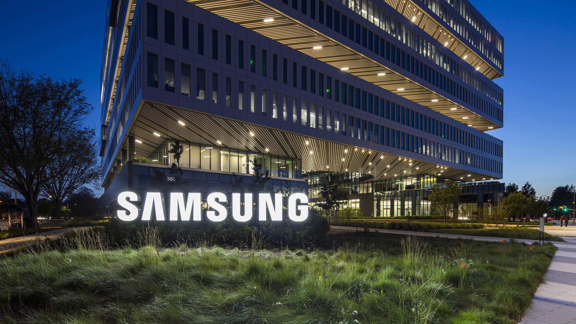 Samsung headquarters building illuminated at dusk with modern glass architecture and glowing Samsung logo outside corporate campus.