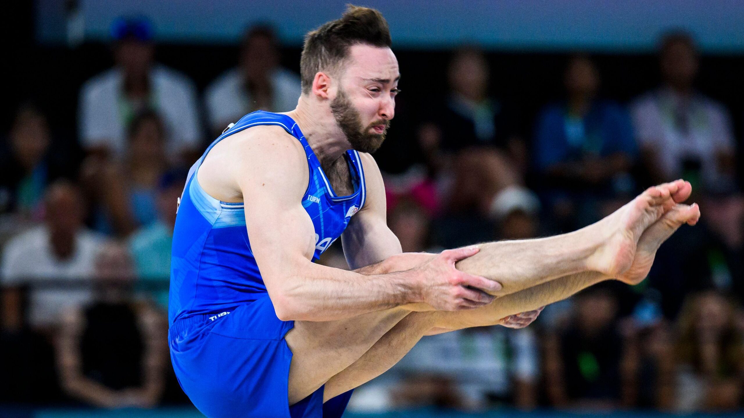 Male artistic gymnast in blue uniform performing a tuck position mid-air during a floor routine at a major competition.