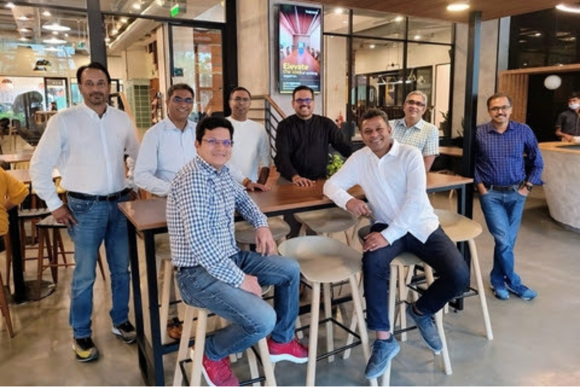Group of seven professionals posing casually around high tables in a modern coworking café-style office, with two seated on stools and others standing in the background.