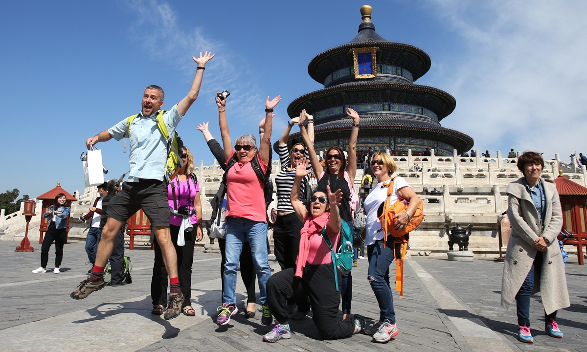 Group of tourists joyfully jumping and posing with raised arms in front of the Temple of Heaven in Beijing on a clear sunny day.