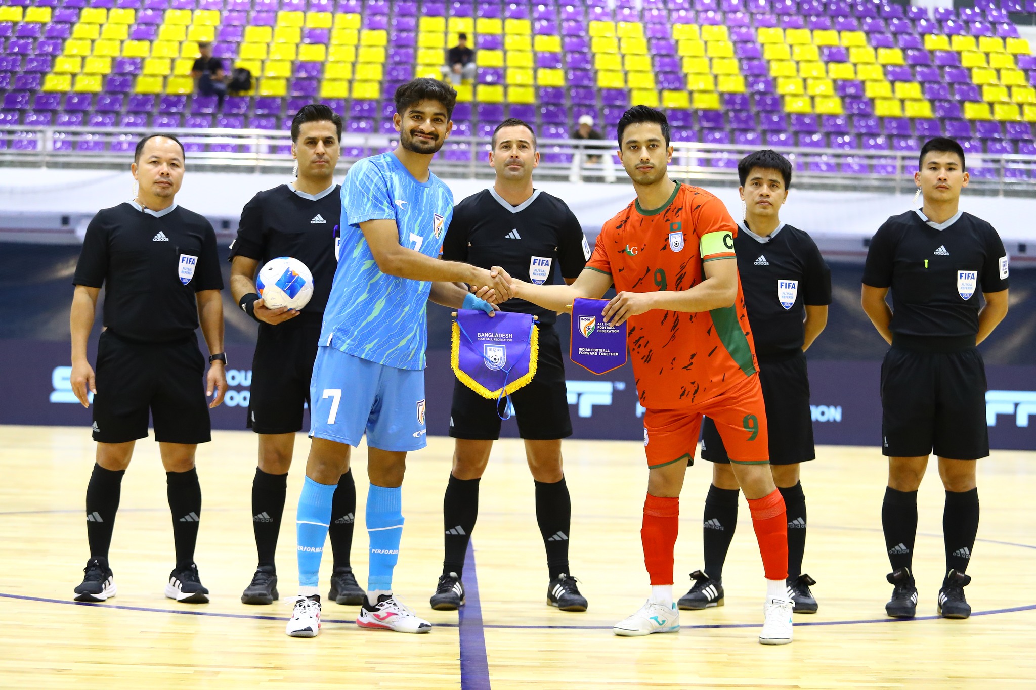 Captains of the India and Bangladesh futsal teams exchange pennants during a pre-match ceremony, flanked by FIFA officials on an indoor court, highlighting regional competition and international futsal action in Asia.