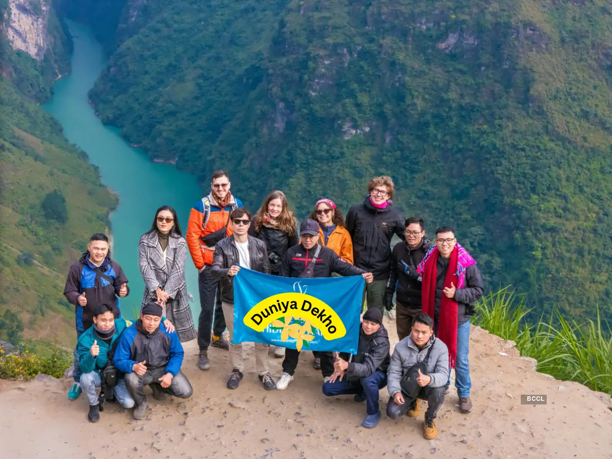 International travel group holding a ‘Duniya Dekho’ flag at a scenic mountain canyon with a turquoise river below.
