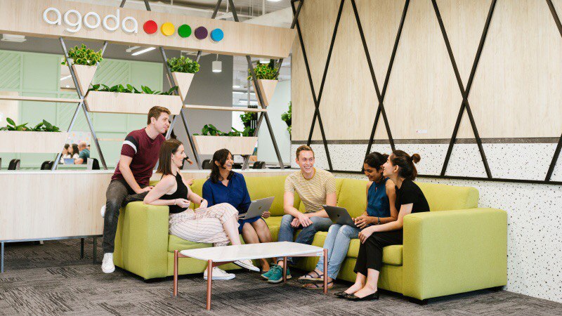 Agoda employees collaborating in a modern open-plan office, with a diverse team seated on green sofas using laptops beneath the Agoda logo, highlighting workplace culture at the global travel technology company.