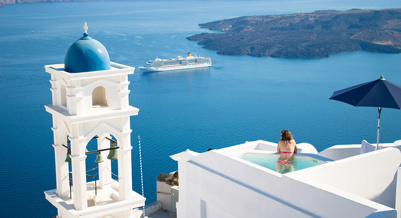 Woman relaxing in infinity pool overlooking Santorini caldera with blue-domed church and cruise ship in the Aegean Sea, Greece.